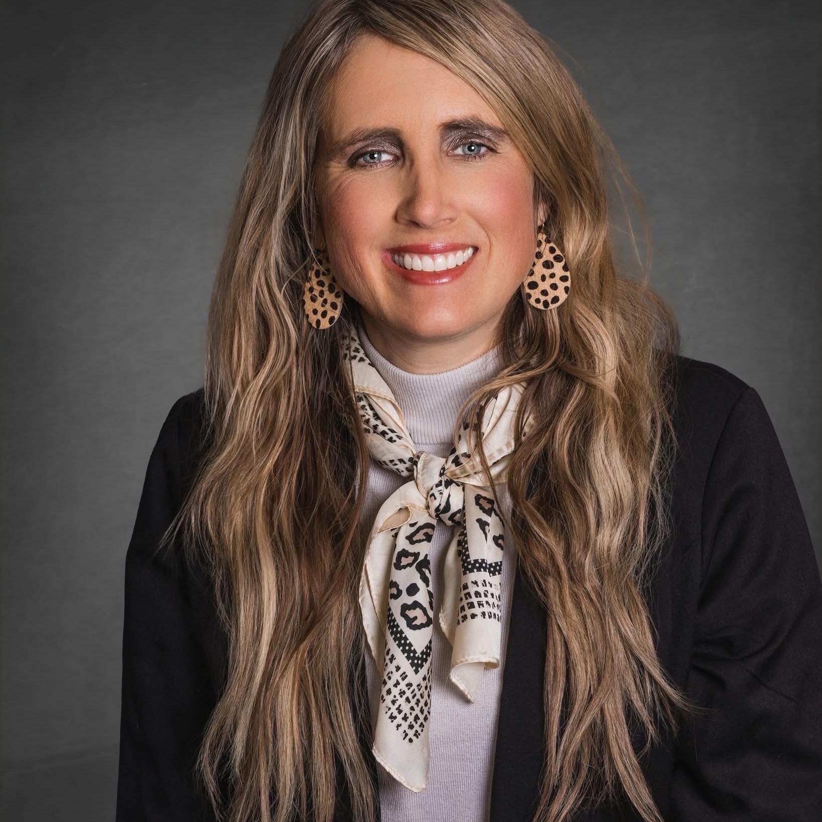 Portrait of Dr. Sarah Diamond, a professional woman with long wavy blonde hair, wearing a patterned scarf and leopard-print earrings, smiling against a neutral gray studio background.
